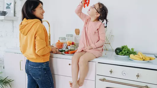 Young girl seated on kitchen counter next to mother looking mindfully at cherry tomato vine.
