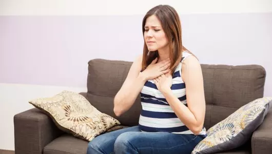 Woman sitting on couch holding her hands to her upper chest in discomfort.