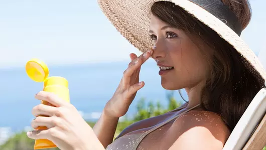 woman sitting outside in a chair with a hat on putting sunscreen on her nose
