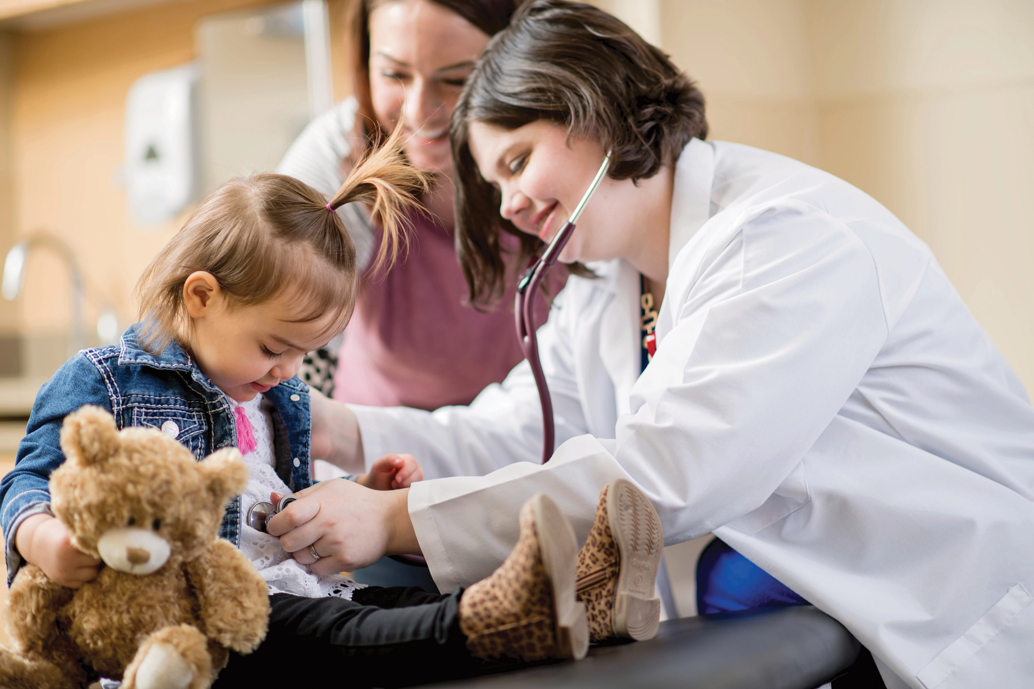 Young daughter getting heartbeat listened to by female doctor while mother looks on.