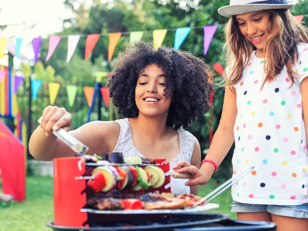 Woman and young girl standing in front of grill with meat and vegetables cooking