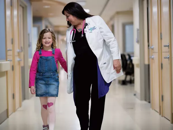 Adult female nurse practitioner walking with young female patient down hospital hallway.
