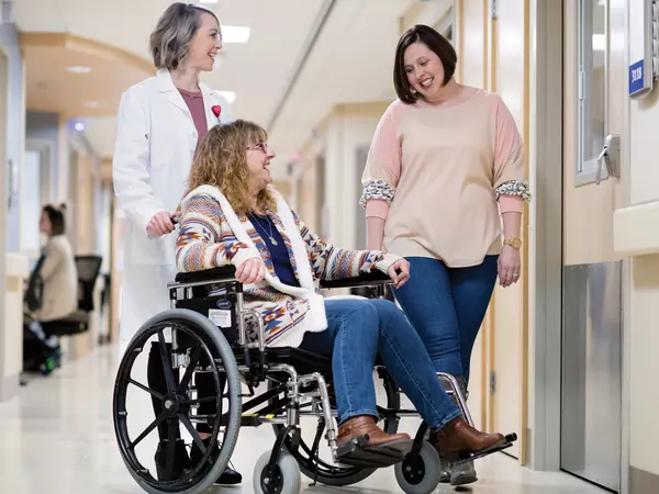 Female adult patient in wheelchair, being pushed by female doctor and adult female friend looking on.