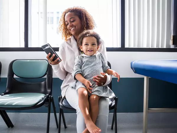 Mother using smart phone while toddler sits on lap at the doctors office.