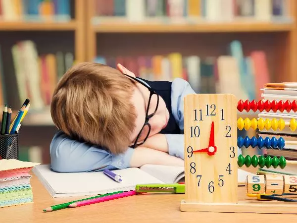 Young boy with glasses is sleeping on his notebook at his desk. In front of him is a cup of pencils, a stack of notecards, and a clock. In the background you can faintly make out a bookshelf full of books.