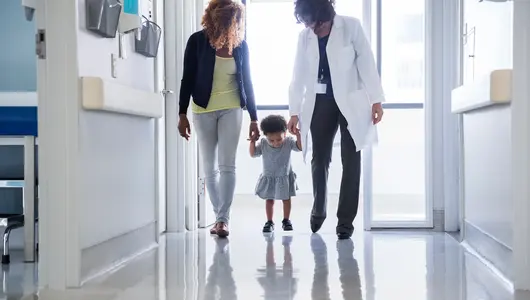 Mother and pediatrician holding hands of toddler while walking in hospital corridor.