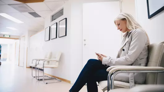 Side view of woman using smart phone while sitting on chair in hospital corridor