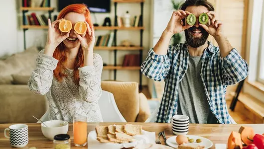 woman and man having fun eating breakfast each holding fruit halves in place of eyes