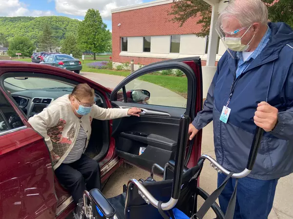 Volunteer helps transport elderly woman from car into wheelchair.