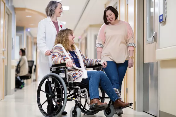 Female adult patient in wheelchair, being pushed by female doctor and adult female friend looking on.